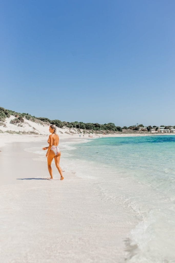A woman walks along a scenic beach shore with clear blue skies and turquoise waters during summer.